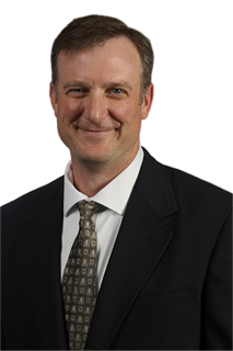 Portrait of a person wearing a black suit jacket, white dress shirt, and patterned gray tie, smiling at the camera against a transparent background.