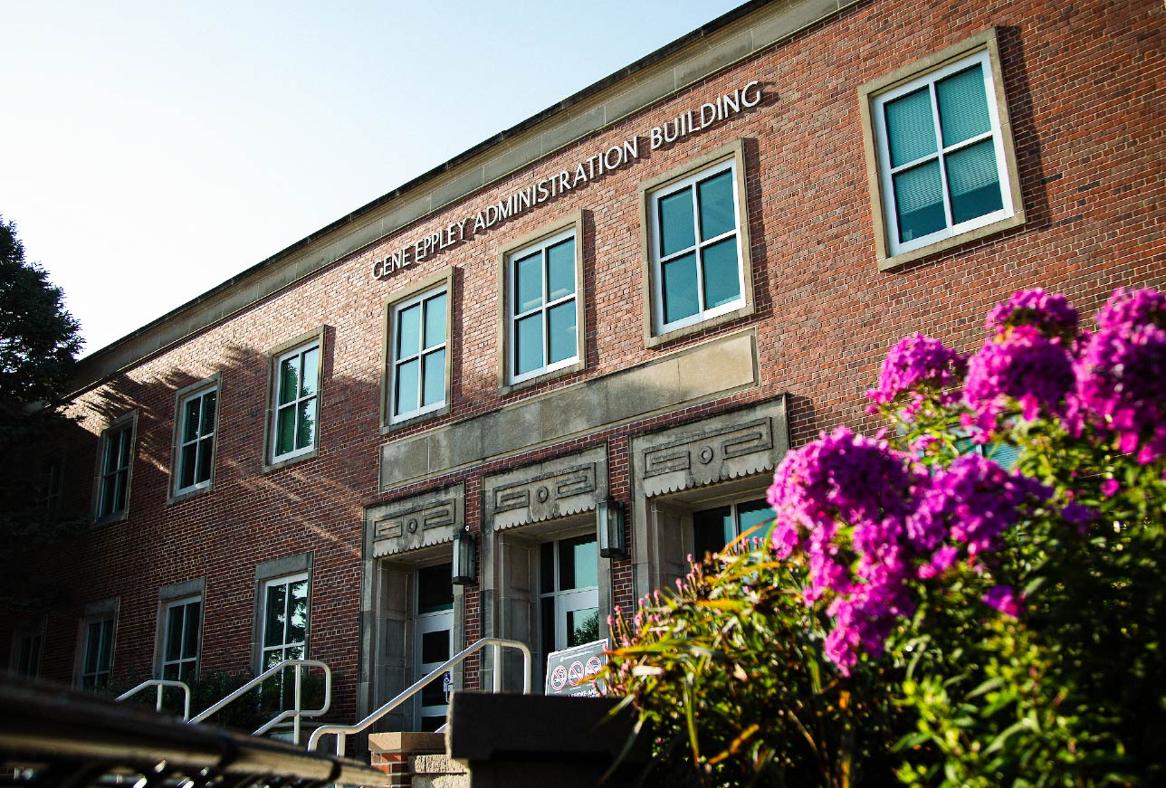 Exterior view of the Gene Eppley Administration Building at the University of Nebraska Omaha, with brick facade, windows, and flowering plants in the foreground.