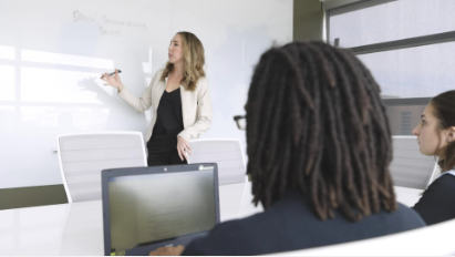 An instructor stands at the front of a classroom, writing on a whiteboard while leading a discussion. Several learners are seated facing the instructor, with a laptop visible in the foreground, creating a collaborative learning and professional development setting.