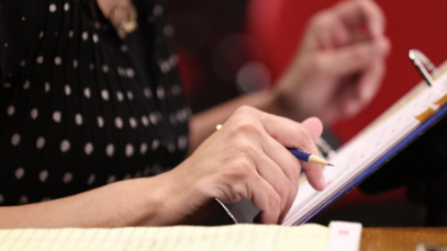 Close-up of a person holding a blue pen and writing on a clipboard or notepad. The image focuses on the person’s hands, with a blurred background and a notebook visible in the foreground, suggesting active note-taking or assessment.
