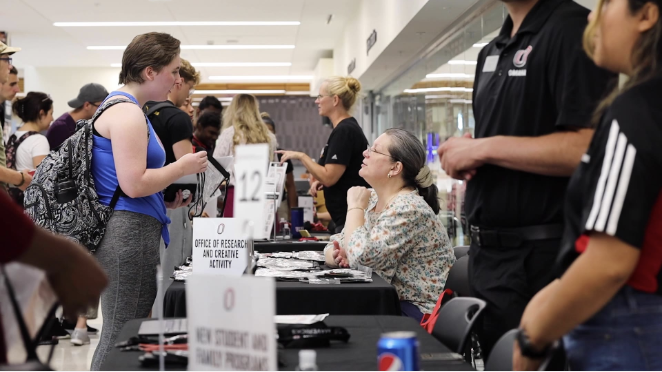 Students gather at an information fair in a campus hallway, speaking with representatives seated at tables. In the foreground, a student with a backpack talks to a staff member at a table marked “Office of Research and Creative Activity,” while other students and exhibitors interact along the busy row of booths.