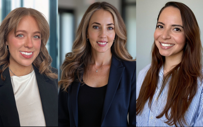 three professional women in business attire smiling in a well-lit office environment.