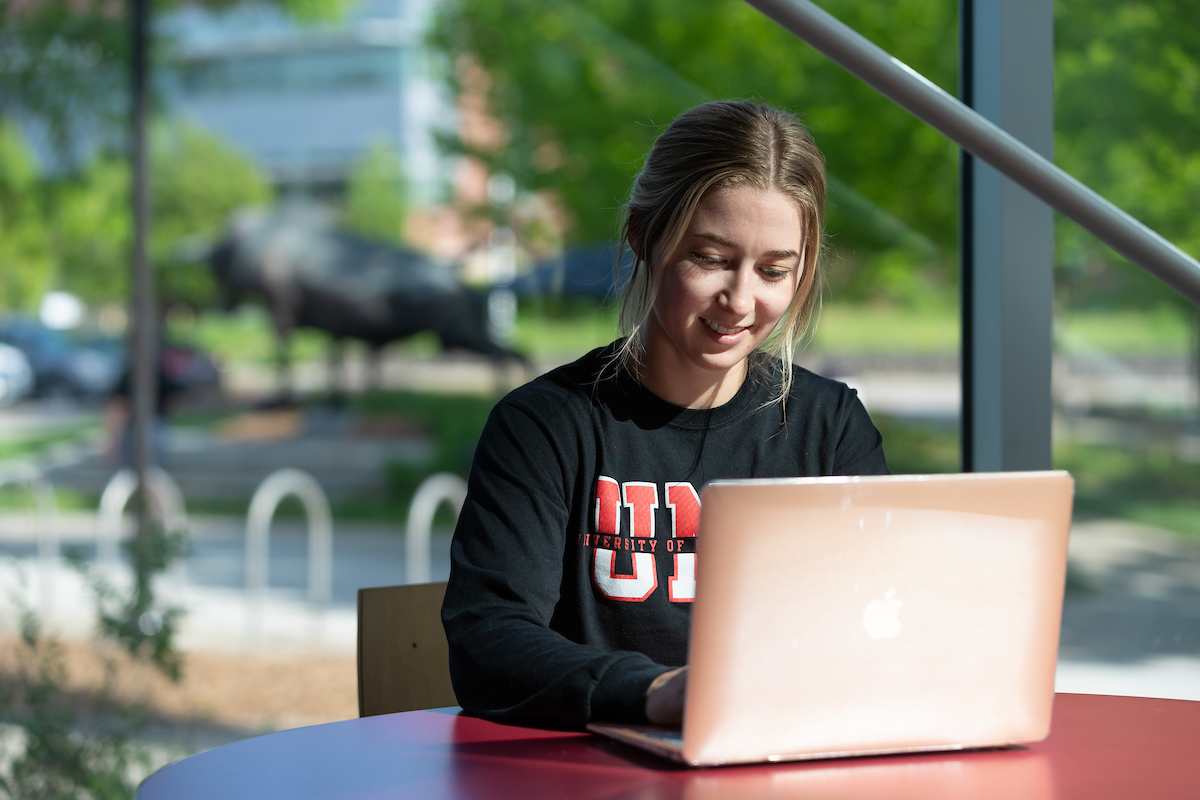 A young woman in a UNO shirt works on her laptop.