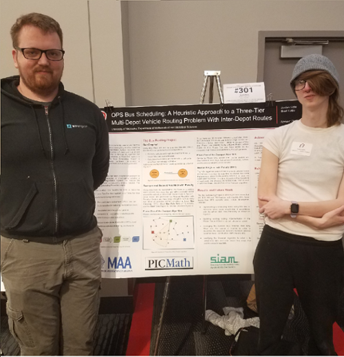 Brad Tuttle (a white man with short brown hair, wearing thin glasses and a black hoodie) and Jordan Sahs (a white woman with medium-length brown hair, wearing wide glasses, a gray beanie, and white t-shirt) stand in front of their poster.