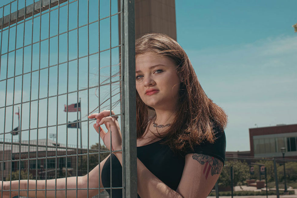 Molly Ashford leans against a chain link fence, facing forward and making eye contact