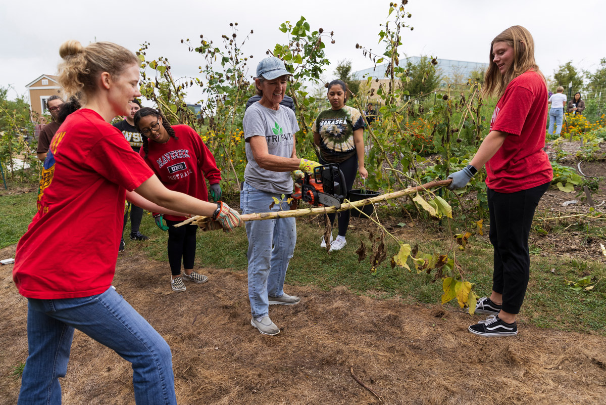 Two women hold the ends of a long branch while a third woman in the enter cuts through it with a chainsaw.