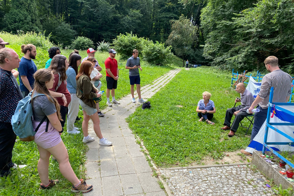 Outisde, an elderly man sits in a folding chair to the right while a group of students to the left talk with him.