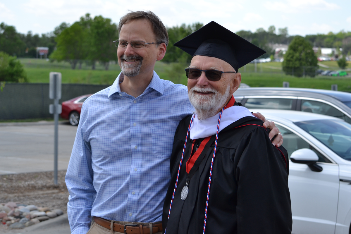 Two men stand together, smiling. One is in a button up shirt and the other is in a cap and gown. 
