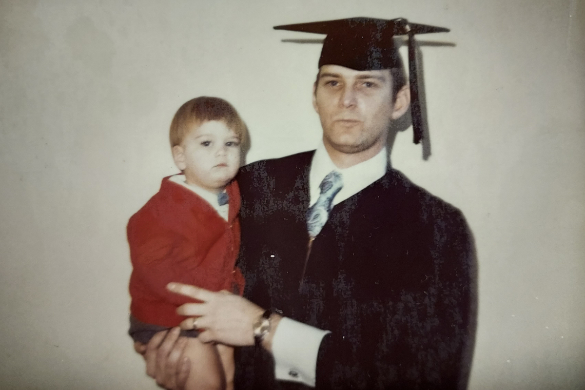 A young man in a cap and gown holds a toddler. 
