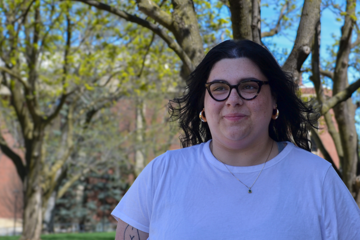 Izzy Vargas stands in front of a green tree. She has long dark hair and glasses, and is wearing a white shirt.