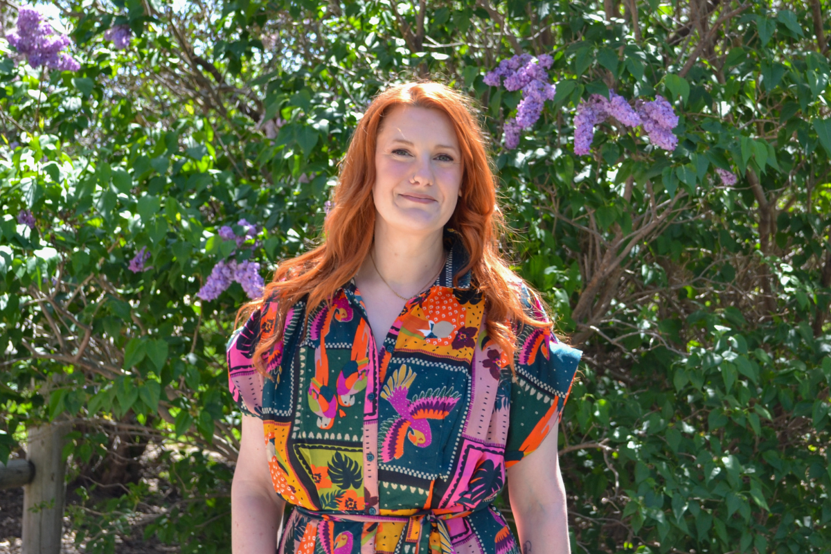 Kelsey Golden stands in front of a tree with purple flowers. She has red hair and a colorful dress. 
