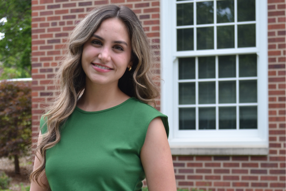 Rebecca wears a green shirt and smiles in front of a brick building.