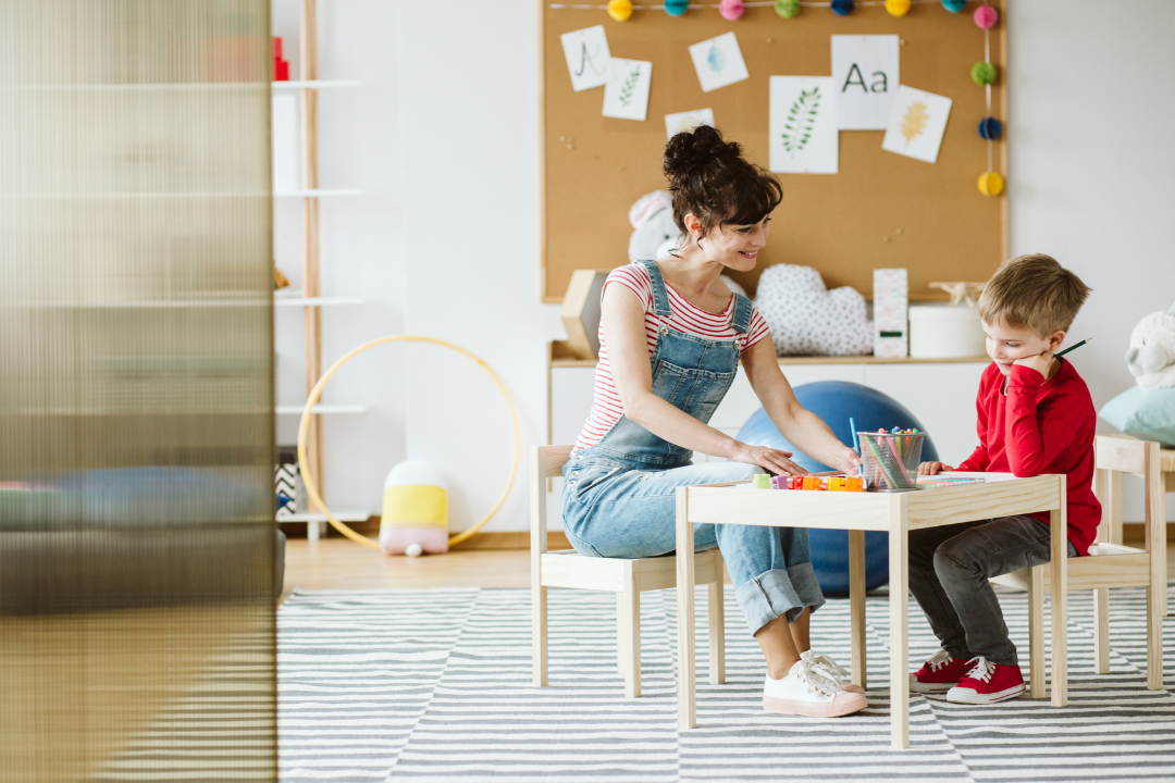 Teacher sits with a young student at a small table, helping with a drawing activity in a bright classroom.
