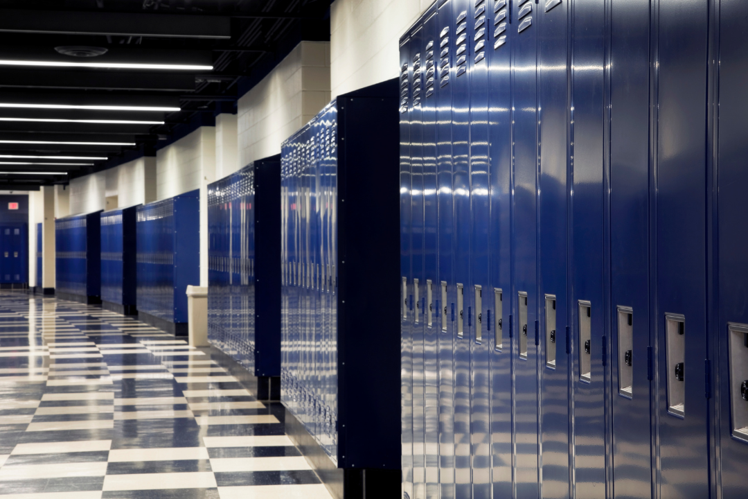 Empty school hallway lined with blue lockers, with a black-and-white checkered floor and overhead fluorescent lighting.