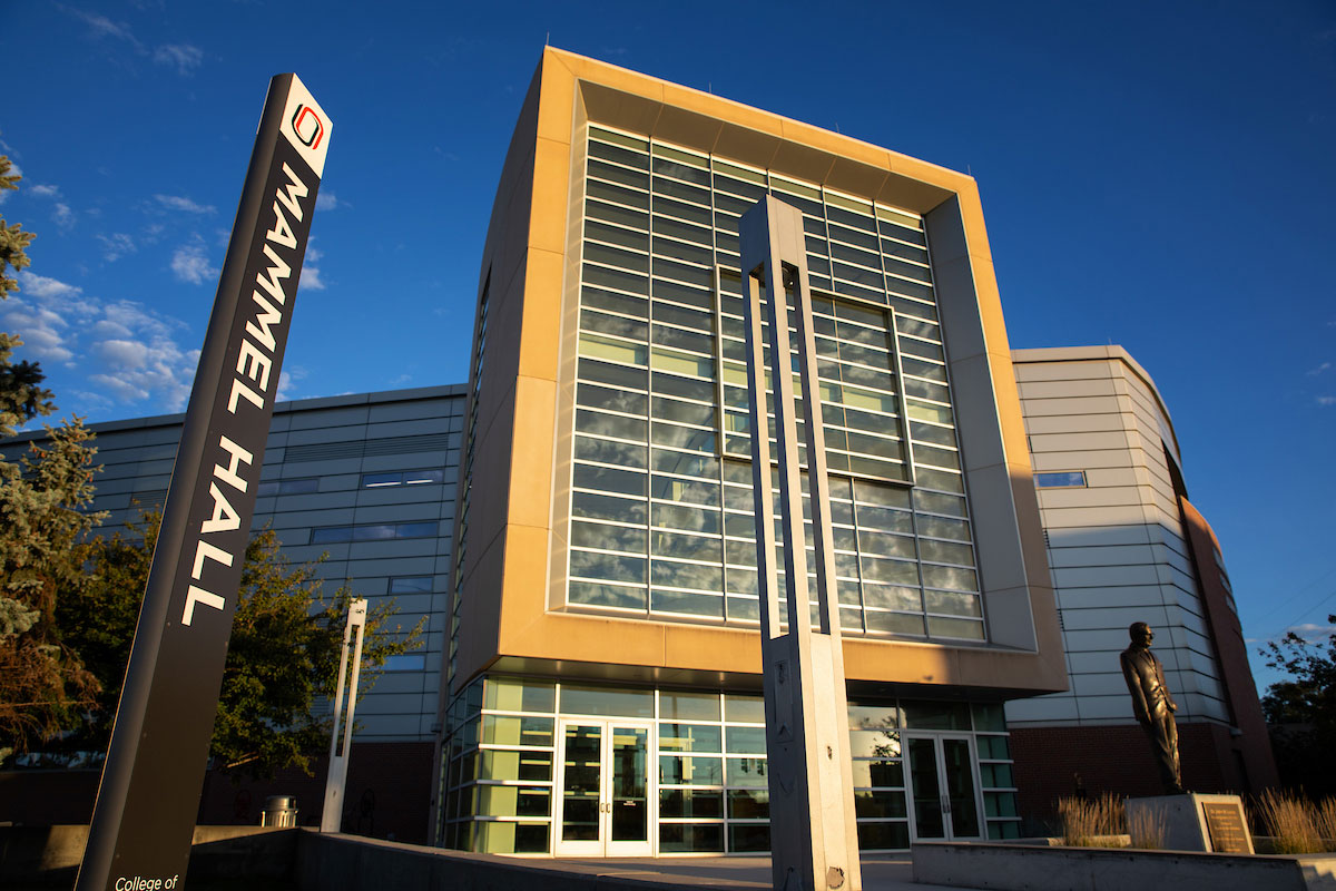 Mammel Hall at the University of Nebraska at Omaha stands under a clear blue sky, with its modern glass facade reflecting clouds. A vertical sign reading “Mammel Hall” is positioned in the foreground, and a statue stands near the building’s entrance.