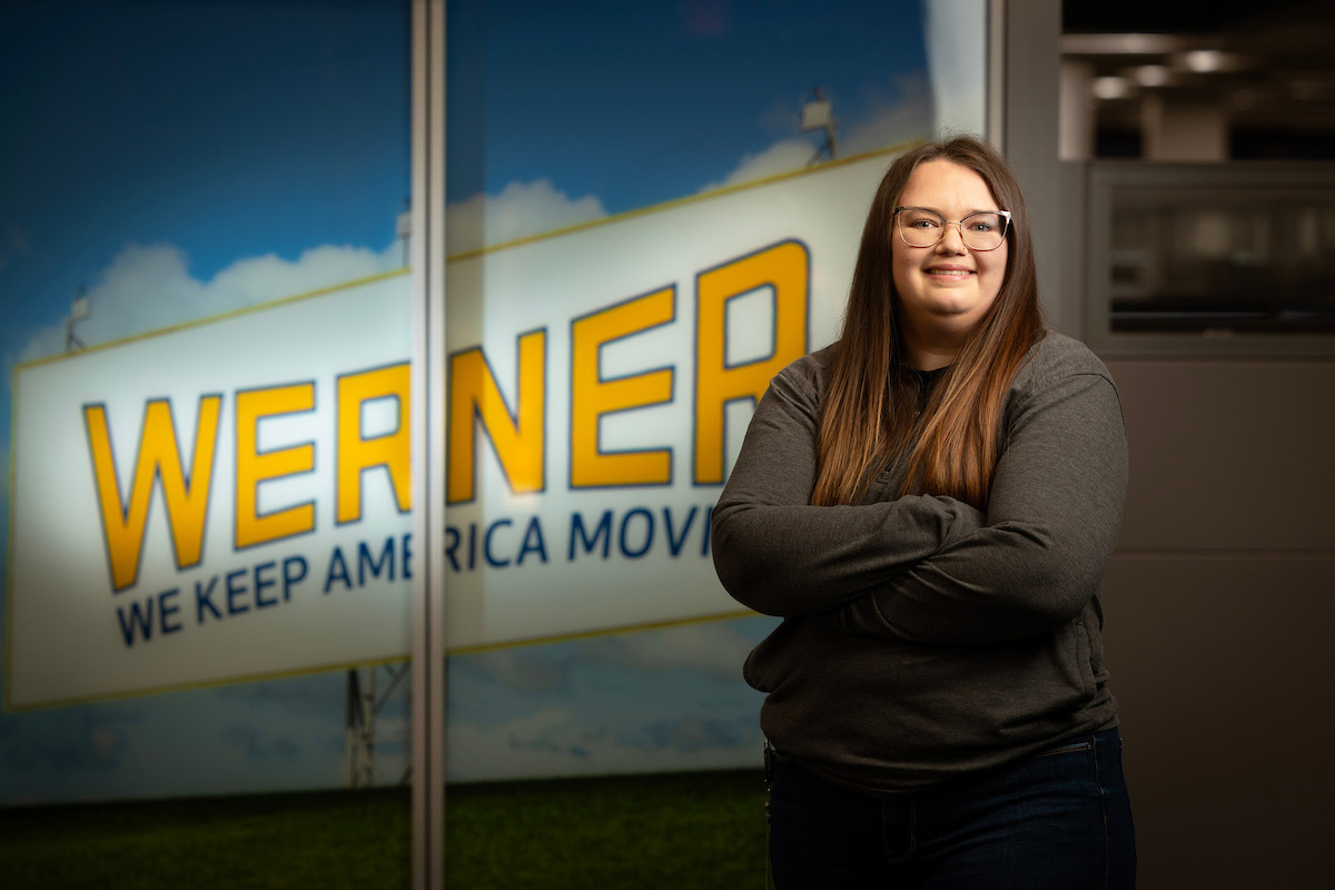 A woman with long hair and glasses stands with her arms crossed, smiling in front of a large Werner sign that reads “We Keep America Moving,” inside an office setting.