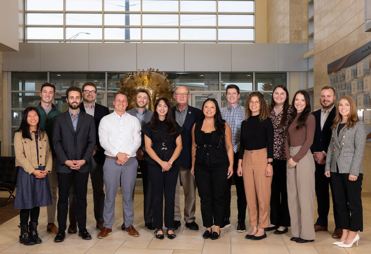 A group of fourteen people stand together indoors, smiling for a group photo in a bright lobby area with large windows and a gold sculpture in the background. The group includes men and women dressed in business or business-casual attire.