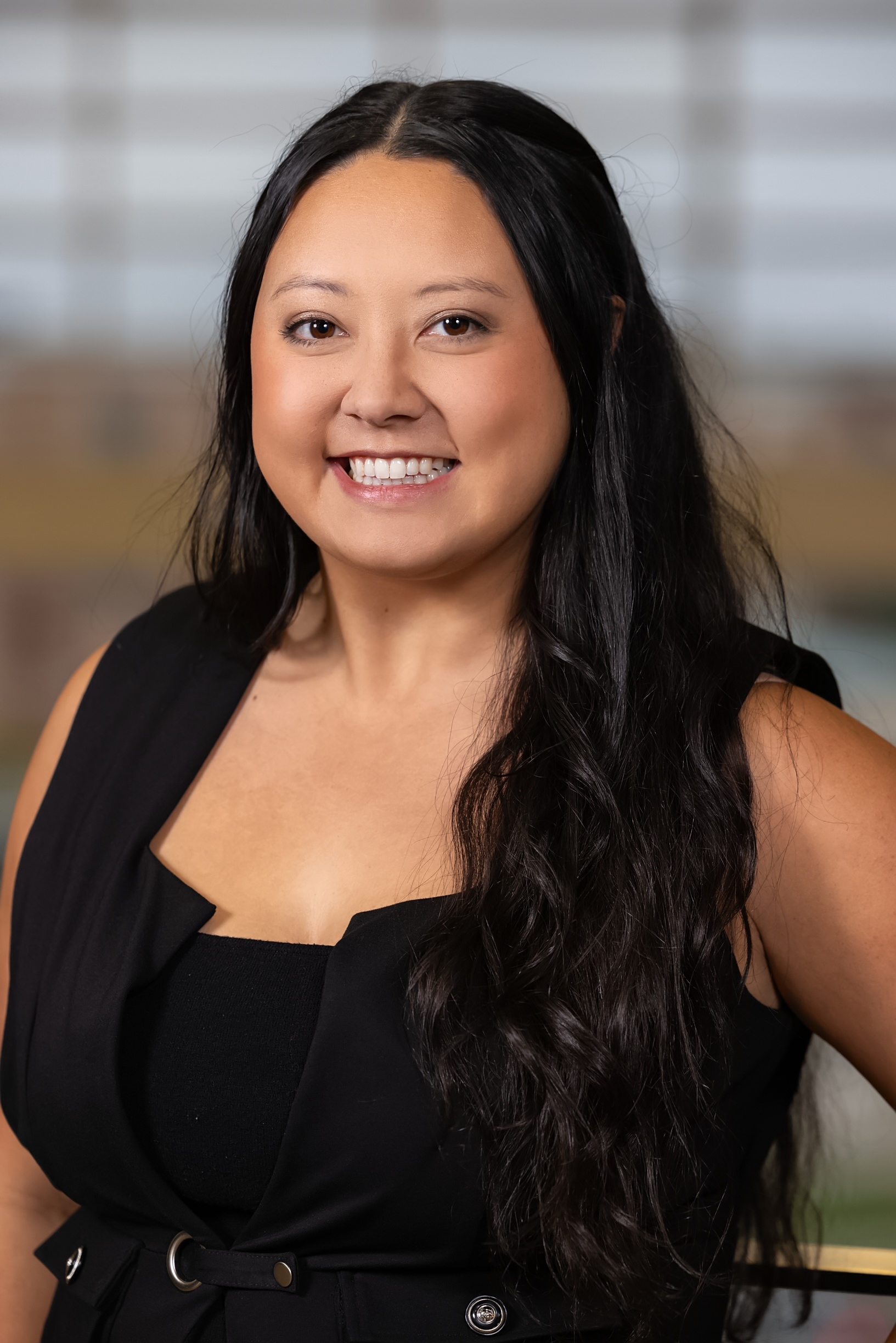 A person with long dark hair smiles at the camera, wearing a sleeveless black top. The background is softly blurred with neutral tones.