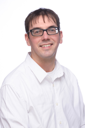 Professional headshot of a person with short dark hair and black rectangular glasses wearing a white button-down shirt. They are smiling at the camera against a plain light gray background.