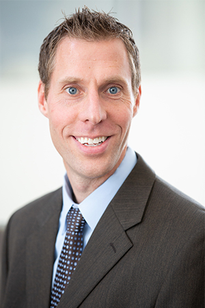 Professional headshot of a person with short light brown hair wearing a dark gray suit, light blue dress shirt, and patterned blue tie. They are smiling at the camera against a softly blurred light gray and blue background.