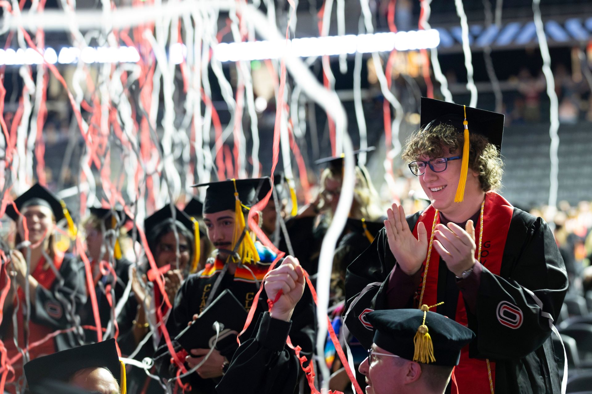 Graduates in black caps and gowns celebrate at a University of Nebraska at Omaha commencement ceremony. Red, white, and black streamers fill the air as one graduate stands clapping and smiling among seated classmates. Excitement and pride are visible across the group.