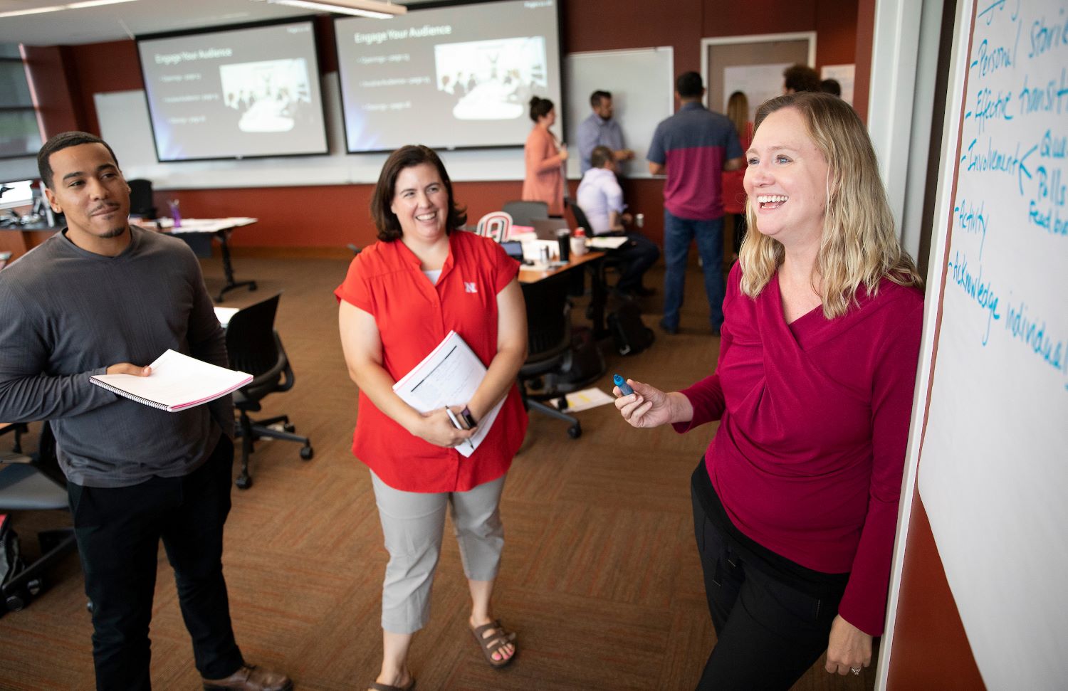 A small group participates in an interactive classroom session, smiling and holding notebooks while a woman stands at a whiteboard writing notes. Presentation slides titled “Engage Your Audience” are displayed on screens in the background, and other participants collaborate in small groups around the room.