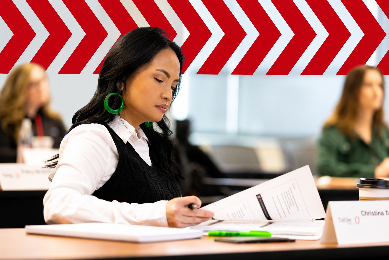 A professional development classroom featuring a participant reviewing course materials at a conference table. A bold band of red chevron arrows spans the upper portion of the image, adding a dynamic graphic overlay while leaving participants unobstructed. Other attendees are softly blurred in the background, creating a focused, modern learning environment.