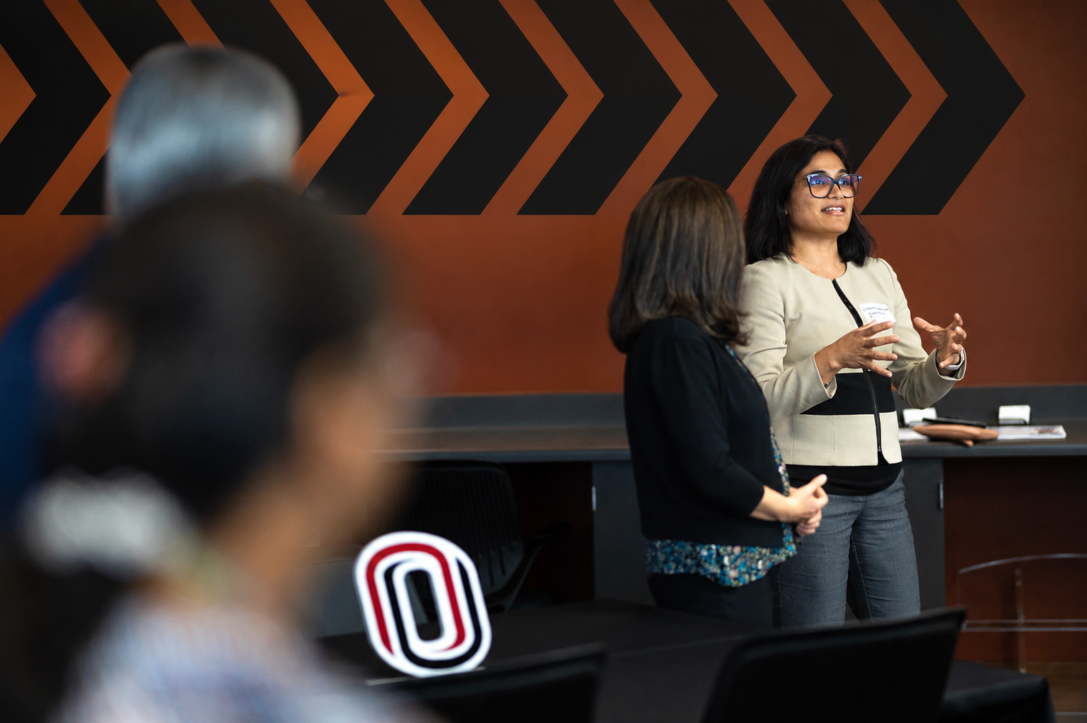 A presenter speaks to an audience at the front of a meeting room while gesturing with both hands, accompanied by another presenter standing nearby. Audience members are visible in the foreground, and a large University of Nebraska at Omaha "O" logo sits on a table. Bold black chevron graphics on an orange wall create a modern backdrop.