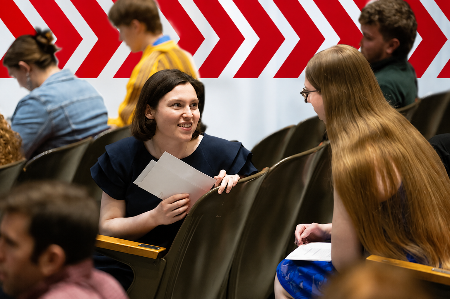 Two students having a conversation in an auditorium, with red chevron graphics in the background.