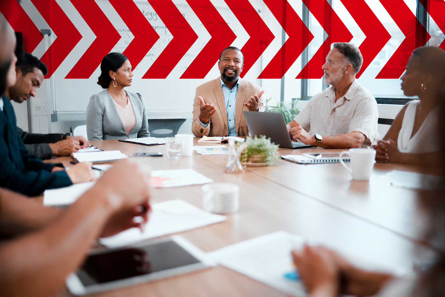 A diverse group of professionals sits around a conference table during a strategy meeting. A person at the center gestures while speaking, as colleagues listen, take notes, and work on laptops. Large red chevron graphics span the glass wall behind them, creating a bold, modern office backdrop.