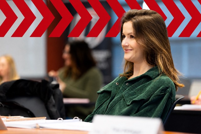 A young professional sits attentively in a classroom during an Executive MBA session, with notebooks and course materials on the table in front of her. A bold red chevron graphic spans the upper portion of the image, adding a dynamic visual element while other participants and the classroom background remain softly blurred.