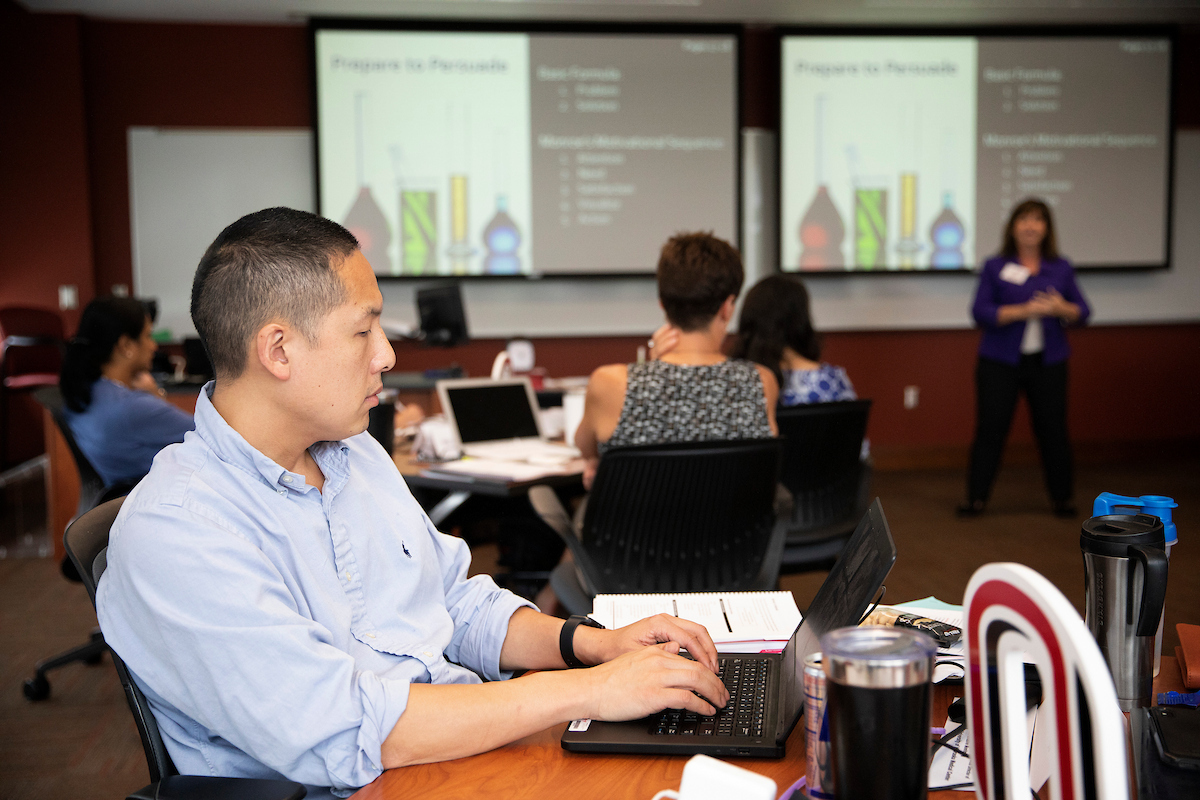 A man sits at a table in a classroom, typing on a laptop while taking notes, as a presenter stands at the front of the room. Other participants sit nearby, and presentation slides are displayed on large screens behind the speaker.