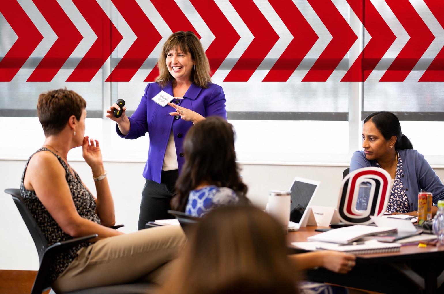 A presenter in a purple blazer leads an interactive workshop, gesturing while speaking to participants seated around a conference table. Attendees listen, discuss, and use laptops and notebooks. A University of Nebraska at Omaha "O" logo sits on the table, with bold red chevron graphics across the glass wall creating a modern meeting-room backdrop.