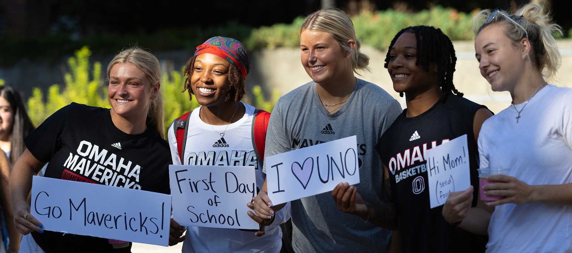 Five smiling students, wearing Omaha Mavericks shirts, stand together outdoors holding handwritten signs that read: 'Go Mavericks!', 'First Day of School', 'I ♥ UNO', and 'Hi Mom! (send food)'. It appears to be a sunny day on campus.
