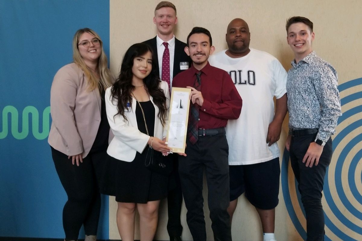 6 students are lined up in a row and smiling, and hold an award between them. They stand in front of a backdrop that says American Marketing Association Omaha.