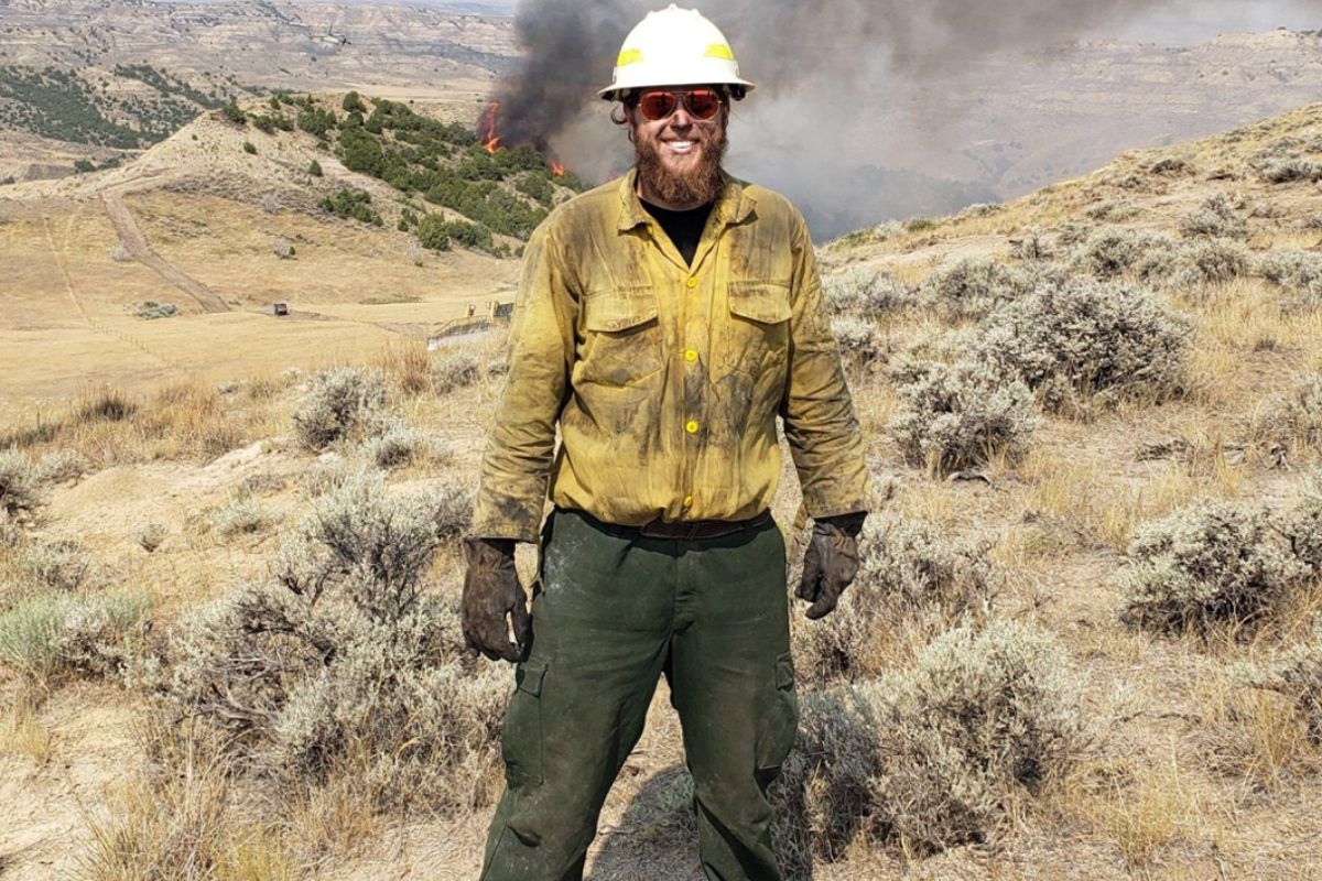 An image of a man in firefighting gear standing in front of a prairie. A fire burns in the distance.