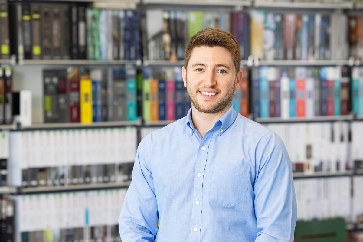 A man in a blue shirt stands in front of a bookcase and smiles at the camera