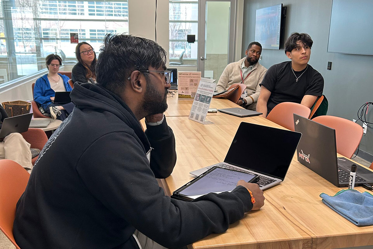 Several people are seated around a large wooden table in a modern meeting room. In the foreground, a person in a black hoodie is working on a laptop and writing on a tablet. Others sit further back, facing a wall-mounted screen, and appear to be listening attentively. Laptops, notebooks, and a microfiber cloth are on the table. The room has large windows with natural light and a ceiling-mounted camera above the table.