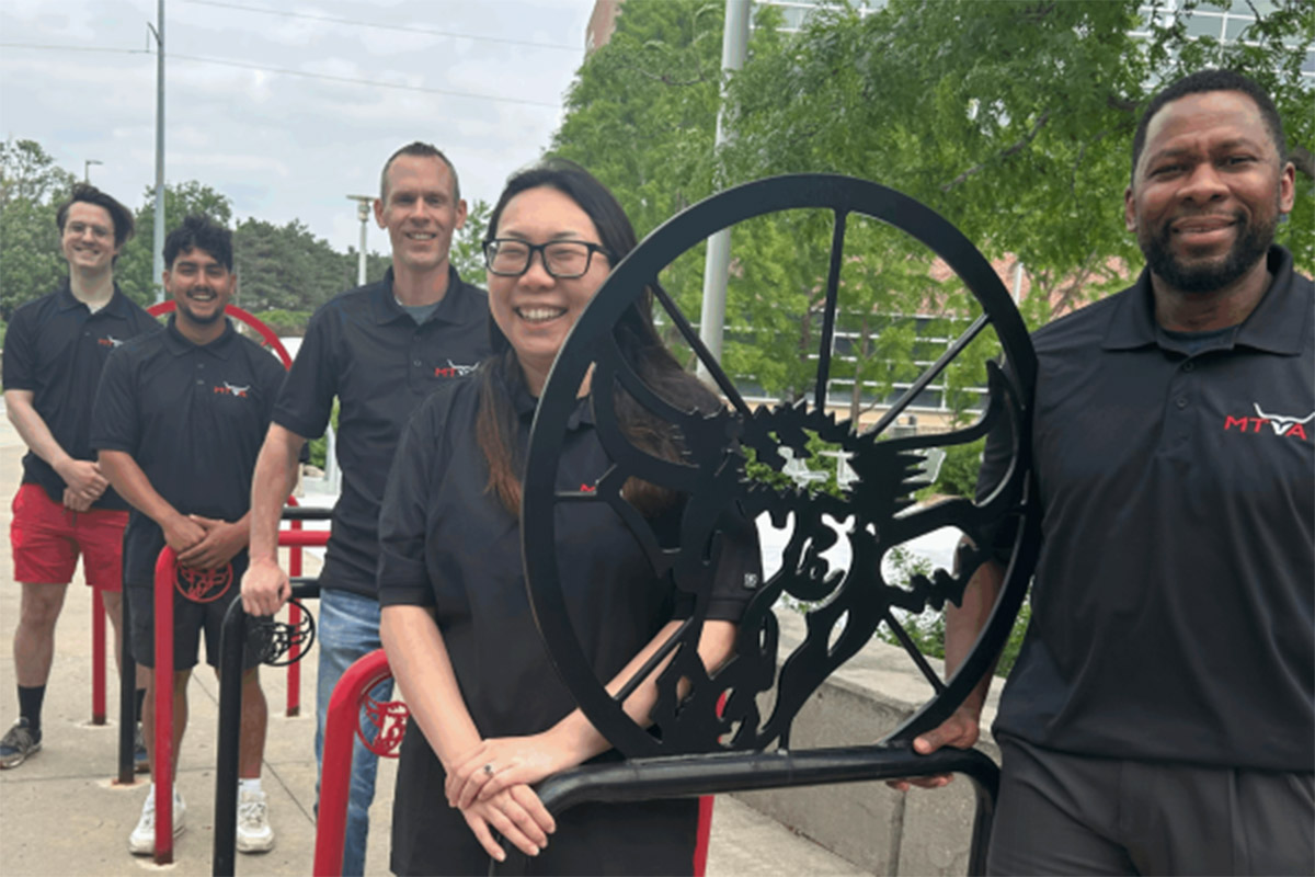Five people wearing matching black polo shirts with the MTVA logo stand outdoors in front of decorative metal bike racks. Two people in the front are smiling and holding onto the racks, while the others stand in a line behind them. Trees and a building are visible in the background.