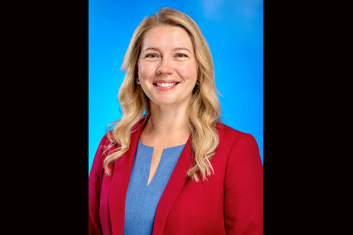A smiling woman with long blonde hair wearing a red blazer and blue blouse, standing in front of a bright blue background.