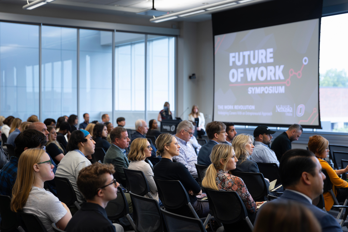 "A conference room filled with attendees listening to a presentation at the ‘Future of Work Symposium.’ The audience is seated in rows of chairs facing a large projection screen that displays the event title. Two presenters stand near a podium in front of the room.