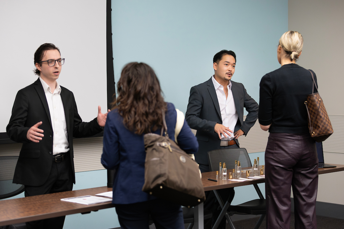 wo men in business suits speak with two women after a presentation. They are standing by a table with water bottles in a conference room.
