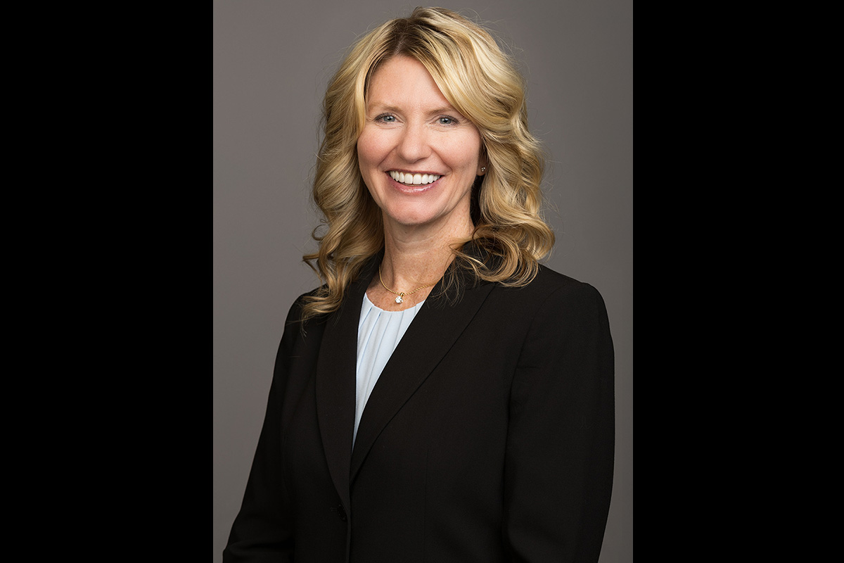 Professional headshot of a smiling woman with shoulder-length wavy blonde hair, wearing a black blazer over a light-colored blouse and a small necklace. She is facing the camera against a plain gray background.