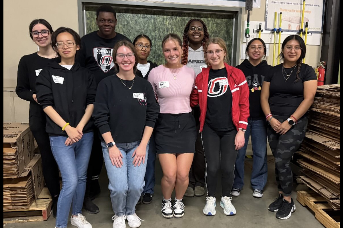 A group of ten students pose together in a warehouse setting, smiling for the camera. They are standing in front of stacked cardboard boxes and a sign that says ‘Thank you for your support.’ Most are wearing casual clothes and University of Nebraska at Omaha gear.