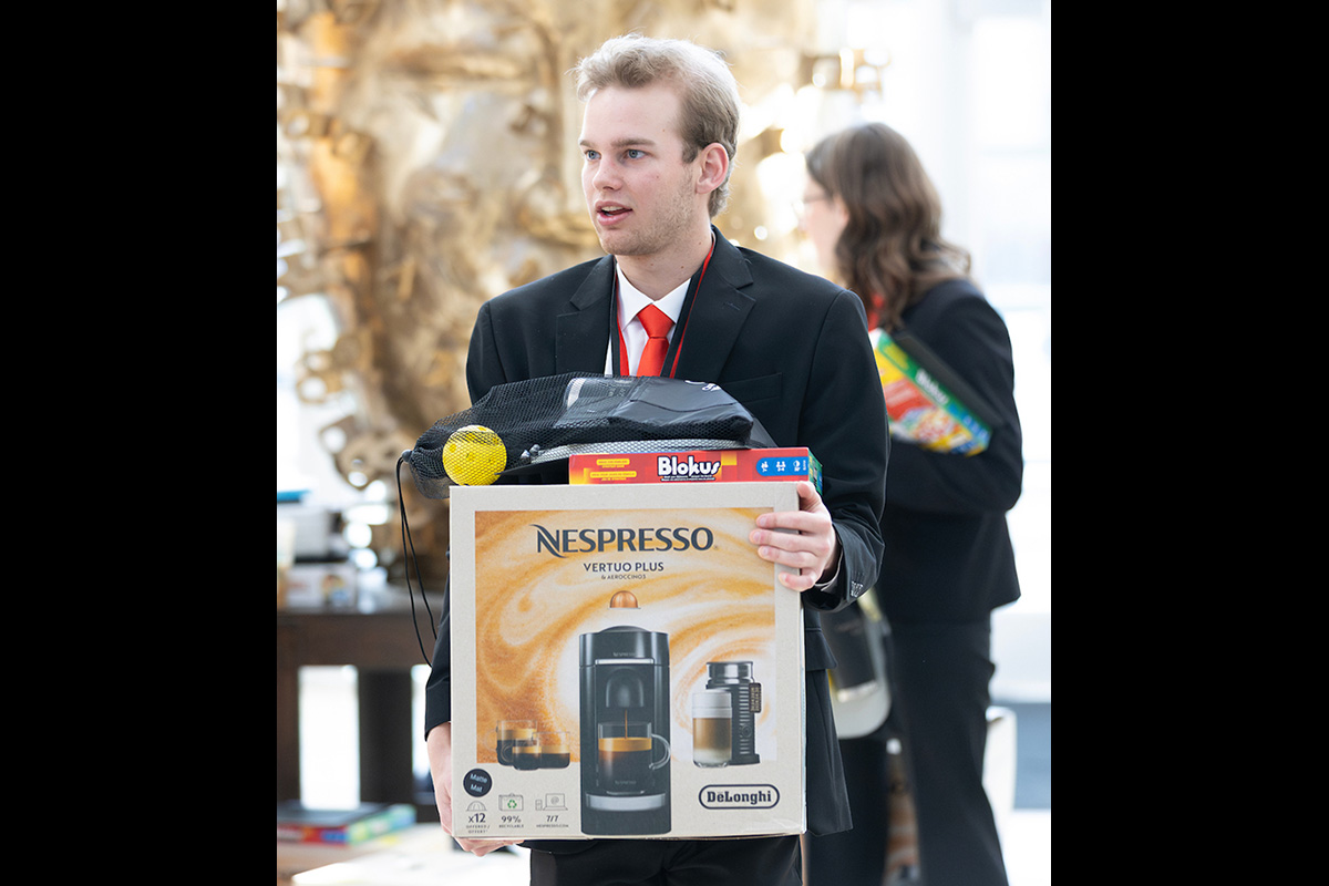 A young man in a suit and red tie carries a large Nespresso Vertuo Plus coffee maker box along with other prizes, including a Blokus game and a mesh bag containing a yellow ball. He appears to be at an event or awards gathering, with people and displays visible in the background.