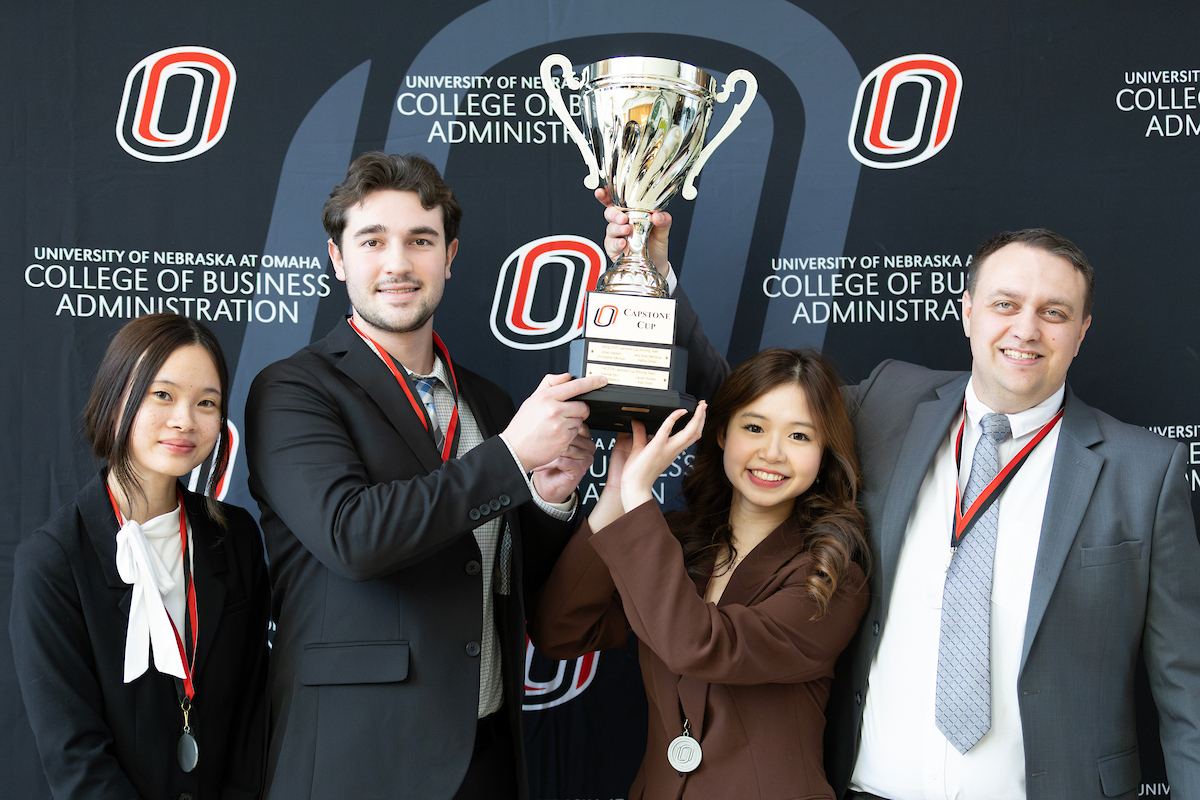 Four people stand smiling in front of a University of Nebraska at Omaha College of Business Administration backdrop. They hold up a large trophy labeled “Capstone Cup,” wearing medals around their necks as they pose together in celebration.