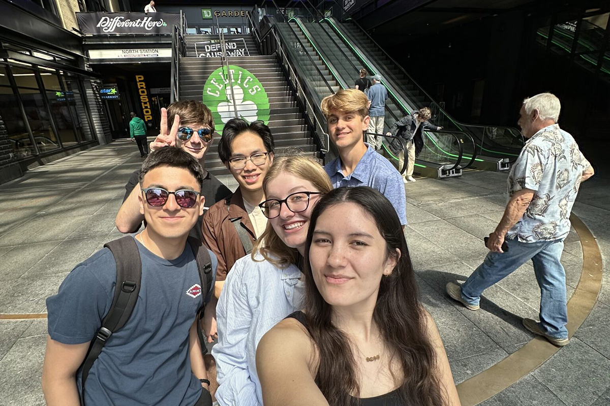 A group of seven young adults take a close-up group selfie outdoors in a large, modern plaza. They are smiling and standing close together. Behind them are wide stairs and escalators leading upward, with green accents and a large circular sign that reads “Celtics” with a shamrock symbol. Storefronts and signage surround the area. A few other people are visible in the background walking through the space. The lighting is bright daylight, and the overall mood is upbeat and social.