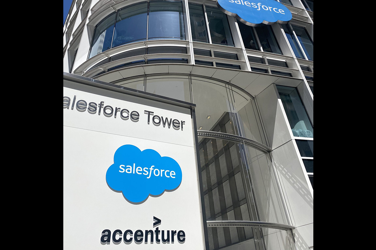A close-up view of the exterior of a modern office building labeled “Salesforce Tower.” In the foreground is a white sign featuring the blue Salesforce cloud logo with the word “salesforce” inside it, and the Accenture logo below. Behind the sign, the building’s curved glass façade and multiple floors are visible, reflecting daylight. The image is taken from a low angle, emphasizing the height and sleek, contemporary design of the building.