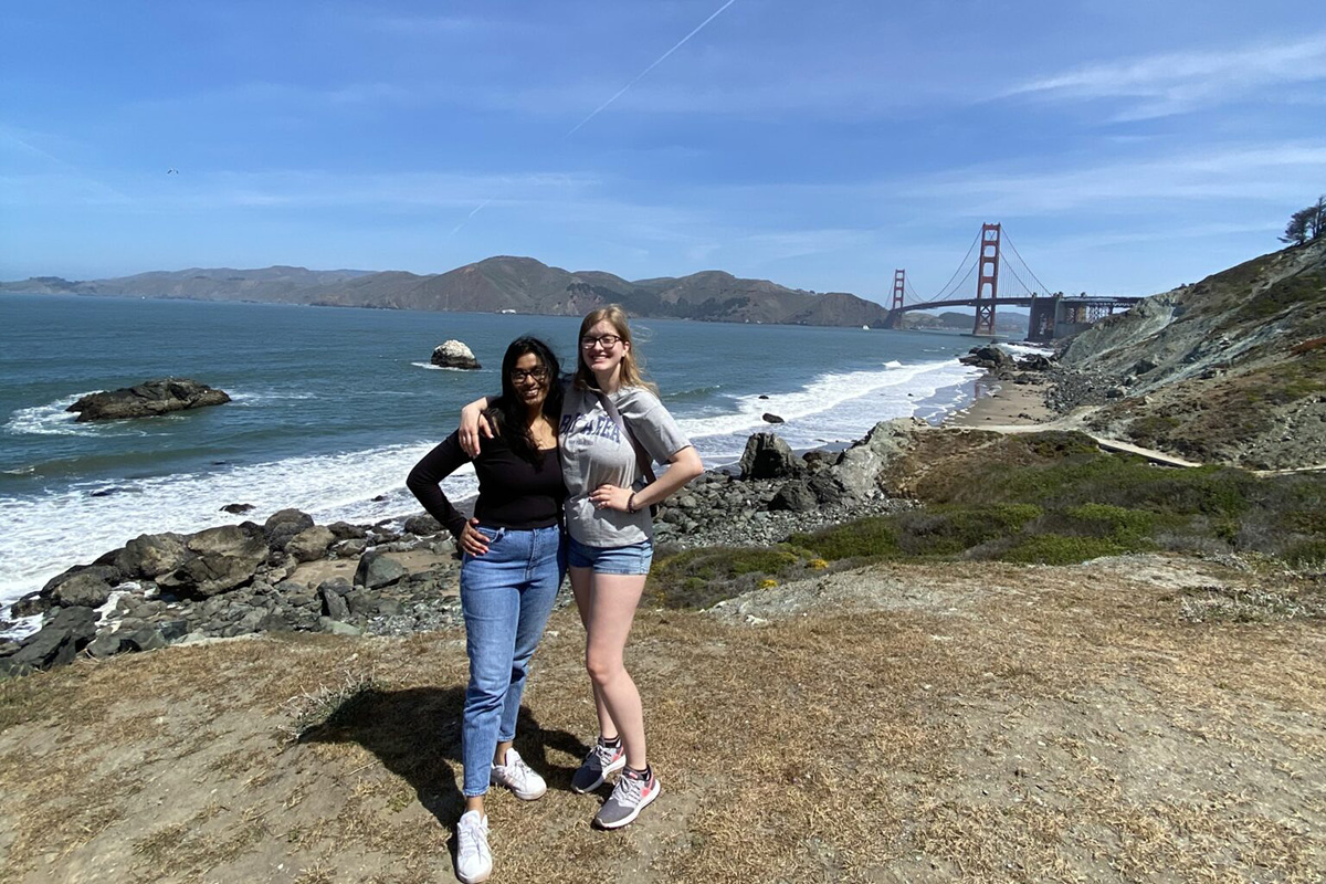 Two young women stand together on a coastal overlook, posing with their arms around each other. They are smiling and dressed casually in jeans, shorts, t-shirts, and sneakers. Behind them is a rocky shoreline with waves breaking along the coast, and the ocean stretches into the distance. The Golden Gate Bridge is visible in the background to the right, partially framed by coastal hills. The sky is clear and blue, and the scene feels bright, breezy, and scenic.
