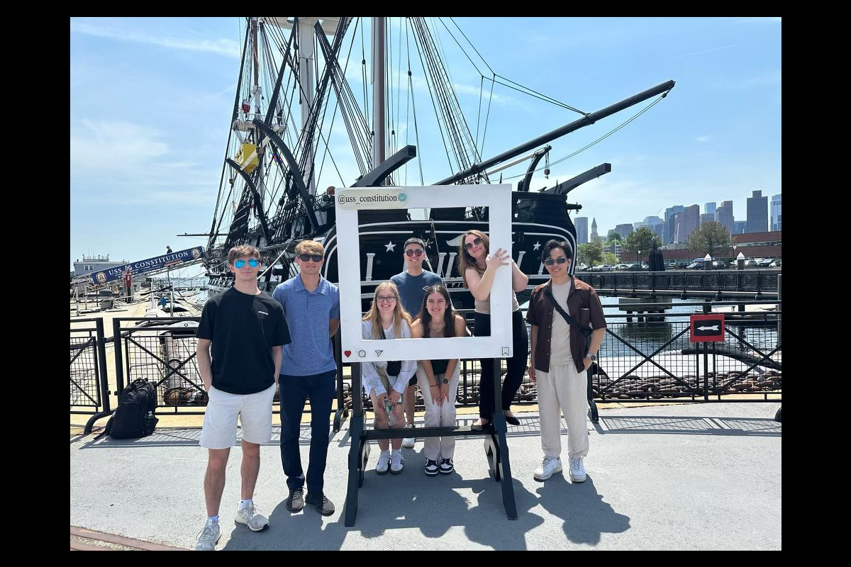 A group of seven young adults pose outdoors at a waterfront museum, standing in front of the historic ship USS Constitution. Some stand on either side of a large, freestanding white frame designed to resemble a social media photo post, while others pose inside it, smiling. The ship’s tall masts, rigging, and dark hull fill much of the background. A city skyline and harbor are visible behind the ship. Everyone is dressed casually, and the scene is brightly lit by sunlight, giving it a fun, tourist-spot feel.